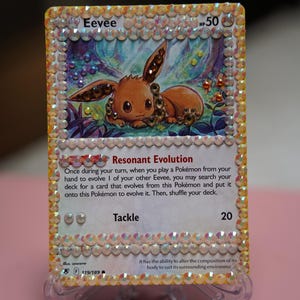 May include: A rectangular Eevee trading card featuring an illustration of the brown Pokémon character, decorated with iridescent rhinestones along the entire border and on the character's body. The card displays the text Resonant Evolution and Tackle.