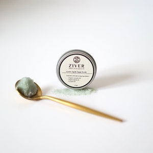 Puede incluir: Una lata plateada con un exfoliante de az&uacute;car de manzana verde en su interior. La lata tiene una etiqueta negra con el texto "Ziver Organiques" y "Green Apple Sugar Scrub". El exfoliante tambi&eacute;n se muestra en una cuchara dorada.
