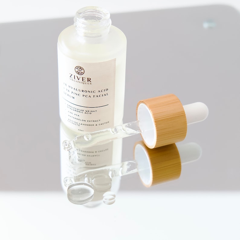 May include: A clear glass bottle with a dropper filled with a serum. The bottle is labeled "Ziver Organiques 2% Hyaluronic Acid 1% Zinc PCA Facial Serum". The dropper is made of bamboo and has a white tip.