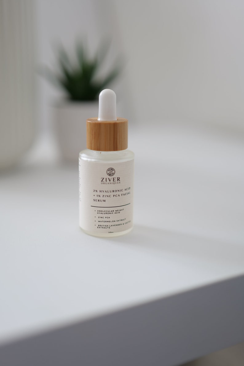 May include: A clear glass bottle with a wooden cap and a dropper top. The bottle is labeled "Ziver Organiques 2% Hyaluronic Acid + 1% Zinc PCA Facial Serum". The label also lists the ingredients: "Molecular Weight Hyaluronic Acid, Zinc PCA, Watermelon Extract, British Lavender & Chamomile Extracts".