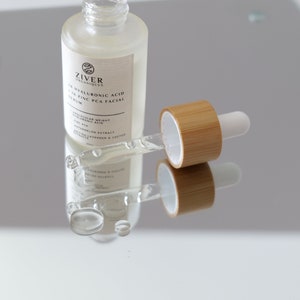 May include: A clear glass bottle with a dropper filled with a serum. The bottle is labeled "Ziver Organics 1% Hyaluronic Acid + 2% Zinc PCA Facial Serum". The dropper is made of bamboo and has a white tip.