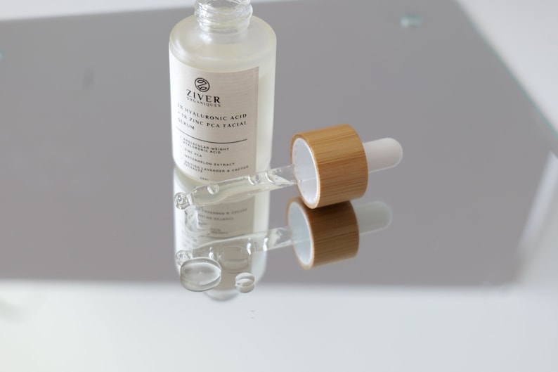 May include: A clear glass bottle with a dropper filled with a serum. The bottle is labeled "Ziver Organics 1% Hyaluronic Acid + 2% Zinc PCA Facial Serum". The dropper is made of bamboo and has a white tip.