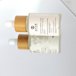 May include: A clear glass bottle with a bamboo dropper top and a white label. The label reads "Ziver Organiques 2% Hyaluronic Acid 1% Zinc PCA Facial Serum". The bottle is sitting on a reflective surface.
