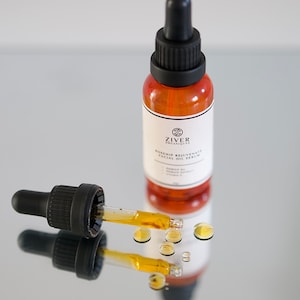 May include: A glass bottle of rosehip facial oil serum with a dropper. The bottle is amber colored and has a black cap. The dropper is filled with a golden oil and there are drops of oil on the surface.