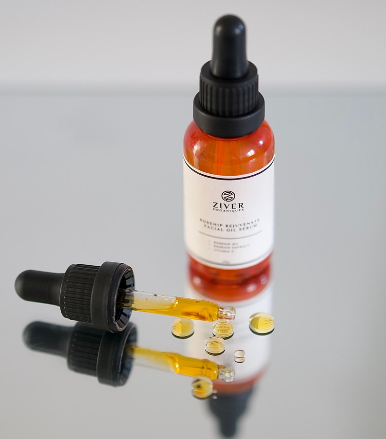 May include: A glass bottle of rosehip facial oil serum with a dropper. The bottle is amber colored and has a black cap. The dropper is filled with a golden oil and there are drops of oil on the surface.