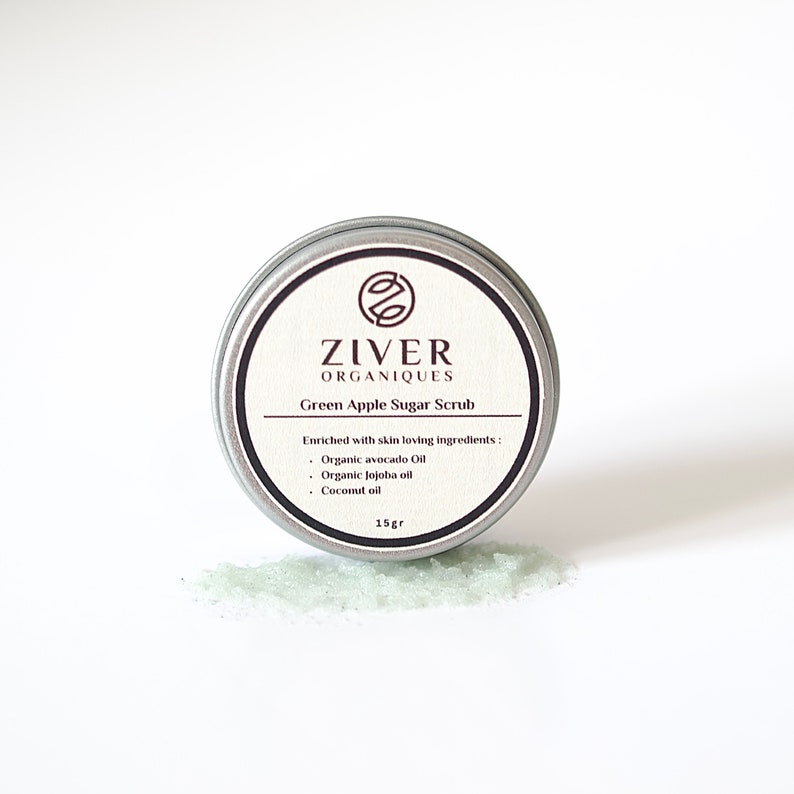Puede incluir: Una lata de plata con una etiqueta blanca que dice "ZIVER ORGANIQUES Green Apple Sugar Scrub" y enumera los ingredientes: "Organic avocado Oil, Organic Jojoba oil, Coconut oil". La lata est&aacute; sobre una superficie blanca con una peque&ntilde;a cantidad de exfoliante de az&uacute;car verde derramado delante de ella.