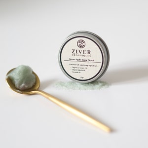 Puede incluir: Una peque&ntilde;a lata plateada con una tapa verde que contiene un exfoliante de az&uacute;car de manzana verde. La lata est&aacute; etiquetada como "ZIVER ORGANIQUES Exfoliante de az&uacute;car de manzana verde" y enumera los ingredientes como "Aceite de aguacate org&aacute;nico, Aceite de jojoba org&aacute;nico, Aceite de coco". El exfoliante tambi&eacute;n se muestra en una cuchara de oro.