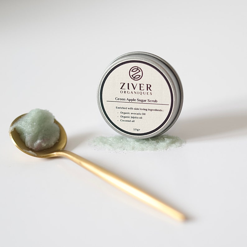 Puede incluir: Una peque&ntilde;a lata plateada con una tapa verde que contiene un exfoliante de az&uacute;car de manzana verde. La lata est&aacute; etiquetada como "ZIVER ORGANIQUES Exfoliante de az&uacute;car de manzana verde" y enumera los ingredientes como "Aceite de aguacate org&aacute;nico, Aceite de jojoba org&aacute;nico, Aceite de coco". El exfoliante tambi&eacute;n se muestra en una cuchara de oro.
