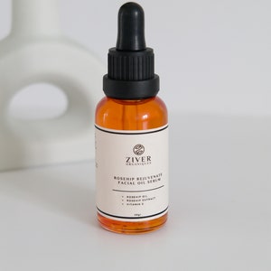 May include: A clear glass bottle with a black dropper top filled with a yellow liquid. The bottle is labeled "Ziver Organiques Rosehip Rejuvenate Facial Oil Serum" and lists the ingredients as "Rosehip Oil, Rosehip Extract, Vitamin E". The bottle is 30 ml.