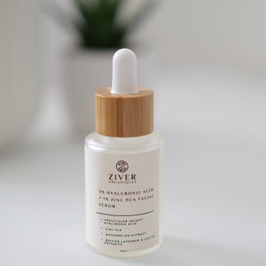 May include: A clear glass bottle with a bamboo lid and a white dropper top. The bottle contains a serum with 2% hyaluronic acid and 1% zinc PCA. The label is white with brown text and the brand name "ZIVER ORGANIQUES".