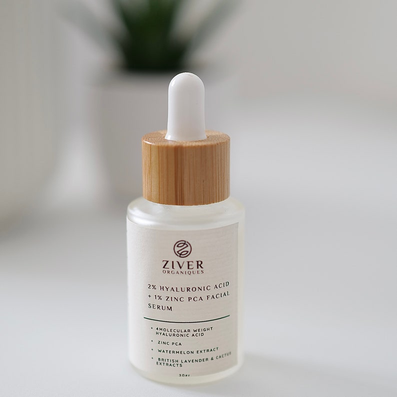 May include: A clear glass bottle with a bamboo lid and a white dropper top. The bottle contains a serum with 2% hyaluronic acid and 1% zinc PCA. The label is white with brown text and the brand name "ZIVER ORGANIQUES".