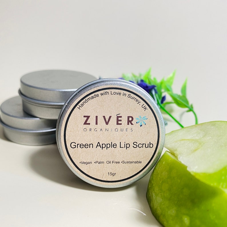 Puede incluir: Una peque&ntilde;a lata de exfoliante labial de manzana verde con el nombre de la marca "ZIV&Eacute;RK ORGANIQUES" y el texto "Handmade with Love in Surrey, UK". La lata est&aacute; sobre una superficie blanca con una rodaja de manzana verde al lado.