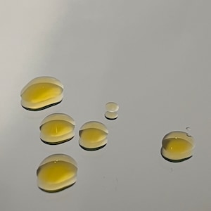 May include: Six drops of a clear, yellow liquid on a white surface.