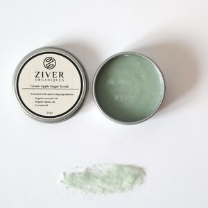 Puede incluir: Un peque&ntilde;o bote de hojalata plateado de exfoliante de az&uacute;car de manzana verde. El bote est&aacute; etiquetado con "ZIVER ORGANIQUES Green Apple Sugar Scrub" y enumera los ingredientes: "Aceite de aguacate org&aacute;nico, aceite de jojoba org&aacute;nico, aceite de coco". El bote est&aacute; abierto y se puede ver una peque&ntilde;a cantidad de exfoliante sobre una superficie blanca.