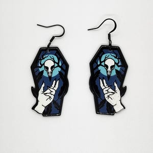 Könnte beinhalten: Ein Paar schwarze, sargförmige Ohrringe. Jeder Ohrring zeigt einen weißen Schädel mit Geweih, eine Hand und einen dunkelblauen Hintergrund mit einer hellblauen Mondsichel. Die Ohrringe haben schwarze Ohrhaken.