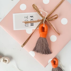 May include: A pink gift box with white polka dots wrapped with a gold ribbon and a tag that says "For You". The box is decorated with a brown and orange tassel.