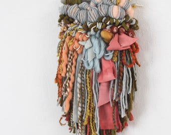 May include: A woven wall hanging with a variety of textures and colors. The piece features strands of orange, pink, and gray yarn, along with blue fabric and a wooden dowel at the top. The artwork is a mix of textures and colors.