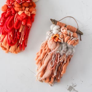 May include: Two handmade woven wall hangings. One is primarily red and orange, with a wooden dowel and hanging string. The other is peach, cream, and gray, with a wooden dowel and string. Both have a textured, layered design.