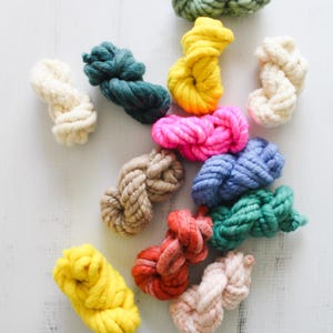 May include: A collection of twelve small, thick yarn skeins in various colors including bright yellow, neon pink, sage green, teal, light blue, cream, beige, soft pink, and variegated red, arranged in a scattered pattern on a white surface.