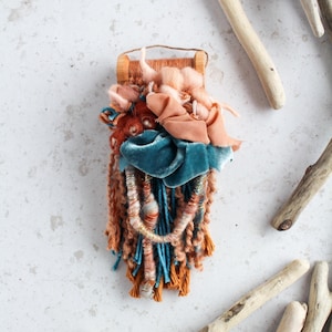 May include: A small, rectangular woven wall hanging with a wooden top. The textile art piece features a mix of textures and colors, including peach, teal, and brown. The design incorporates various yarns, ribbons, and fringe, creating a unique, handcrafted aesthetic.