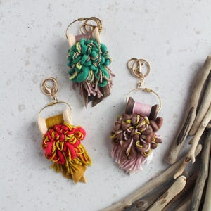 Puede incluir: Tres llaveros hechos a mano con hilo colorido y detalles de madera. Los llaveros presentan herrajes dorados y una variedad de colores de hilo, incluyendo rojo, amarillo, verde y marrón. Los llaveros miden aproximadamente 7.6 cm de largo.