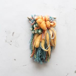 Puede incluir: Un tapiz de pared de arte textil hecho a mano. La pieza presenta una clavija de madera con un grupo de hilo texturizado en tonos de amarillo, verde azulado, naranja y verde. El hilo está dispuesto en un diseño abstracto en capas.