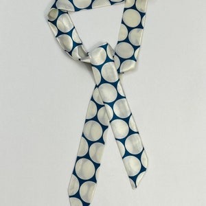 May include: A white satin scarf with a blue polka dot pattern. The scarf is tied in a loose knot.