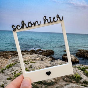 Könnte beinhalten: Ein weißer, quadratischer Fotorahmen mit dem schwarzen Schriftzug "schön hier" am oberen Rand und einem kleinen schwarzen Herzsymbol in der Mitte am unteren Rand. Der Rahmen ist dazu gedacht, vor eine Meeres- und Felsenküstenkulisse gehalten zu werden.