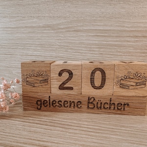 Könnte beinhalten: Holzblockkalender mit der Zahl 20 und dem Schriftzug "gelesene Bücher" darunter. Jeder Block zeigt eine Buchillustration und florale Akzente. Ein Zweig mit rosa Blüten befindet sich im Vordergrund.