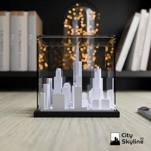 Puede incluir: Maqueta 3D blanca de un horizonte urbano, encerrada en una caja de acr&iacute;lico transparente con base negra. El modelo presenta edificios de diversas formas. El logotipo City Skyline 3D est&aacute; en la esquina inferior derecha.