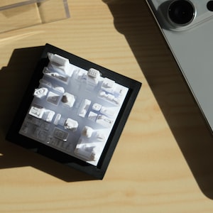 Puede incluir: Una maqueta arquitect&oacute;nica blanca de una ciudad, enmarcada en negro. El modelo muestra formas de edificios y trazados de calles. Un smartphone plateado es parcialmente visible sobre una superficie de madera.