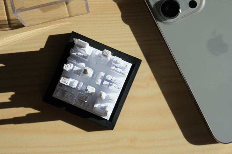 Puede incluir: Una maqueta arquitect&oacute;nica blanca de una ciudad, enmarcada en negro. El modelo muestra formas de edificios y trazados de calles. Un smartphone plateado es parcialmente visible sobre una superficie de madera.