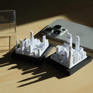 K&ouml;nnte beinhalten: Zwei wei&szlig;e 3D-gedruckte Miniatur-Stadtmodelle auf schwarzen Sockeln, eines in einer durchsichtigen Plastikvitrine. Die Stadtmodelle zeigen hohe Geb&auml;ude und eine Vielzahl kleinerer Strukturen.