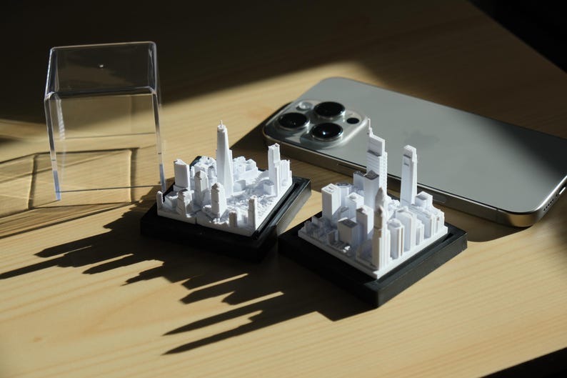 K&ouml;nnte beinhalten: Zwei wei&szlig;e 3D-gedruckte Miniatur-Stadtmodelle auf schwarzen Sockeln, eines in einer durchsichtigen Plastikvitrine. Die Stadtmodelle zeigen hohe Geb&auml;ude und eine Vielzahl kleinerer Strukturen.