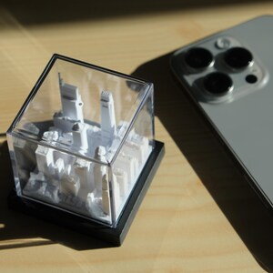 Puede incluir: Una maqueta blanca en miniatura de una ciudad, encerrada en una vitrina transparente en forma de cubo con una base negra. Un smartphone gris con tres lentes de c&aacute;mara est&aacute; al lado de la maqueta sobre una superficie de madera.