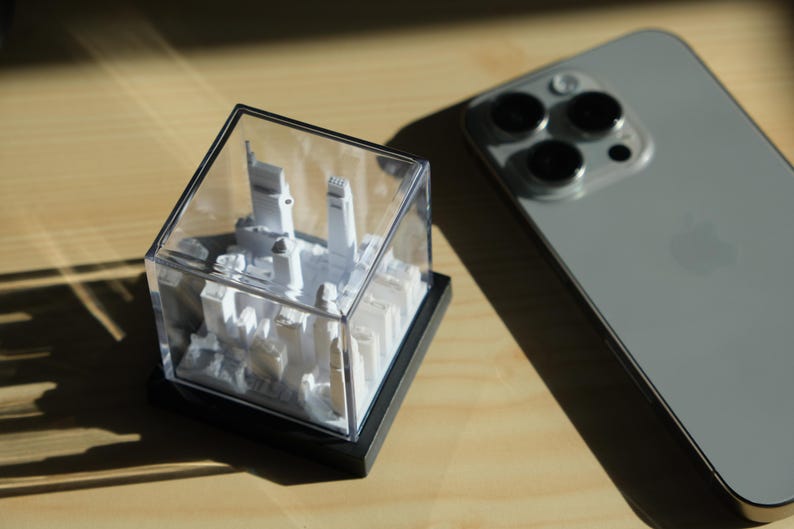 Puede incluir: Una maqueta blanca en miniatura de una ciudad, encerrada en una vitrina transparente en forma de cubo con una base negra. Un smartphone gris con tres lentes de c&aacute;mara est&aacute; al lado de la maqueta sobre una superficie de madera.