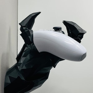 May include: A black geometric deer head wall mount holding a white video game controller. The deer head is mounted on a white wall. The controller is centered in the mount.
