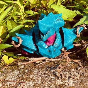Può includere: Una statuetta stampata in 3D di colore blu-verde e viola che rappresenta una creatura con denti e artigli affilati. La statuetta è seduta su un letto di ramoscelli marroni e foglie verdi.