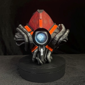 May include: A 3D printed geometric sculpture featuring a central circular eye with a blue glowing light, surrounded by layered dark grey and black mechanical plates, accented with bright orange panels and a small silver rectangular detail.