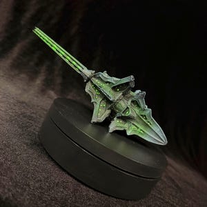 May include: A detailed, green and gray spear-like object with a pointed tip and intricate design. The spear features glowing green accents and is set against a dark background. The object is likely a prop or model.