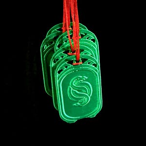 May include: Several green rectangular pendants with a stylized design are suspended from red cords against a black background. The pendants have a carved design and a glossy finish, creating a striking contrast.