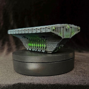 Puede incluir: Modelo gris oscuro de una nave espacial con detalles verdes brillantes. La nave espacial está sobre un soporte giratorio negro, mostrando su intrincado diseño. El modelo tiene un cuerpo plano y rectangular con características detalladas.