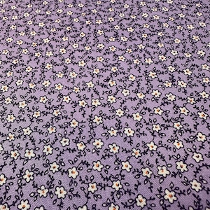 May include: Close-up of a lavender fabric with a repeating floral pattern. Tiny white flowers with orange centers and dark purple stems and leaves cover the surface, creating a delicate, all-over design. The fabric has a soft, textured appearance.