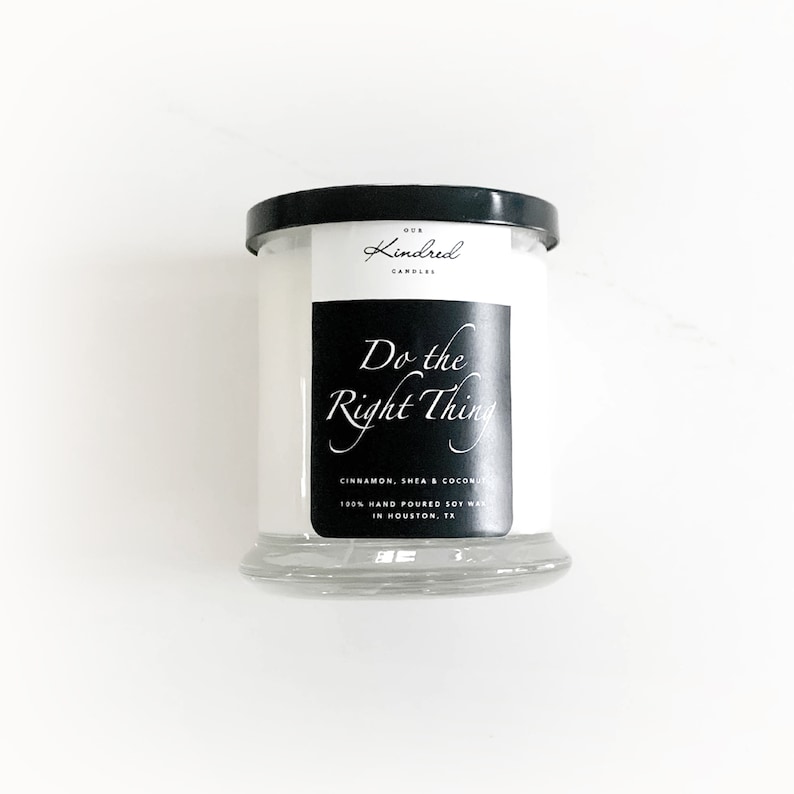 K&ouml;nnte beinhalten: Ein wei&szlig;es Kerzengef&auml;&szlig; mit schwarzem Deckel und Etikett. Das Etikett lautet "Our Kindred Candles" und "Do the Right Thing". Die Kerze ist mit Zimt, Shea und Kokosnuss beduftet. Sie ist handgegossen aus Sojawachs und wird in Houston, Texas, hergestellt.