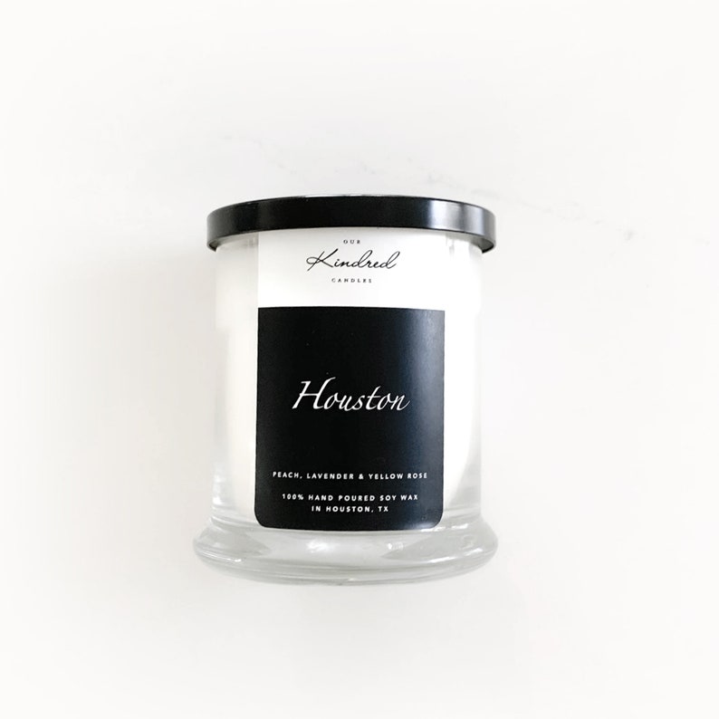Op de afbeelding: Een witte kaars pot met een zwarte deksel en label. Het label zegt "Our Kindred Candles" en "Houston" in een handschrift. De geur is "Perzik, Lavendel & Gele Roos". Het label zegt ook "100% Hand Gegoten Sojawas in Houston, TX."