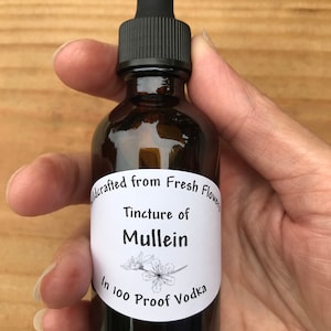 May include: A brown glass bottle with a black dropper top and a white label that reads "Wildcrafted from Fresh Flowers Tincture of Mullein In 100 Proof Vodka".