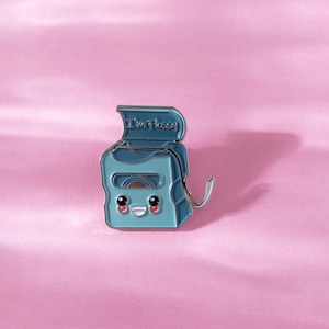 May include: A blue enamel pin shaped like a floss dispenser with a smiling face and the text "I'm Flossy".
