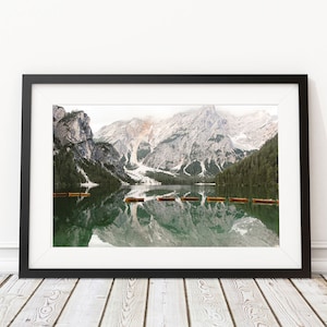 May include: A panoramic view of a mountain lake with snow-capped peaks reflected in the still water. Several wooden boats are lined up on the surface of the lake.