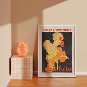 May include: A white framed poster with a black background featuring a woman in a yellow and orange dress dancing. The text "Folies-Berg&egrave;re" and "La Lo&iuml;e Fuller" is printed in red on the poster.