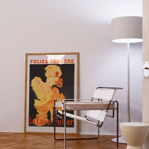 May include: A white leather and chrome chair sits in a room with a wood floor. A framed poster with the text "Folies-Berg&egrave;re La Lo&iuml;e Fuller" is on the wall behind the chair. A white floor lamp with a white shade is in the corner of the room.